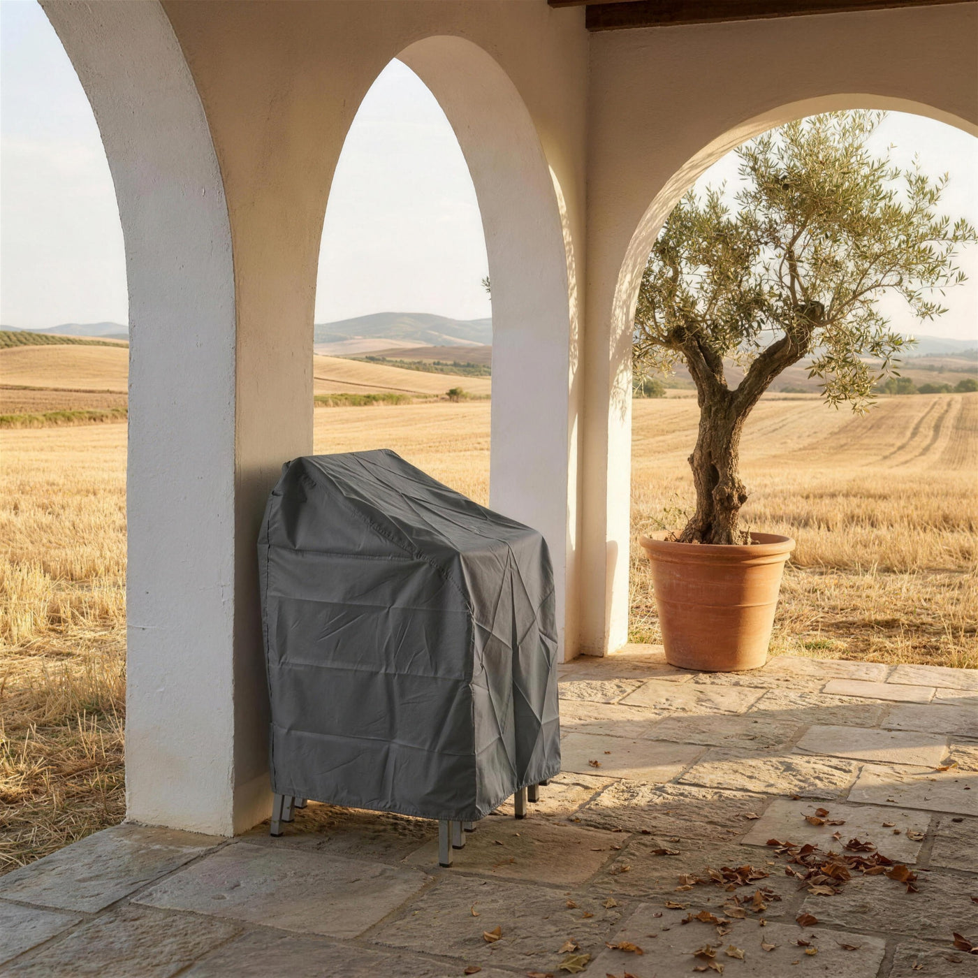 Sillas apiladas y cubiertas con su funda protectora bajo los arcos encalados del porche de un cortijo sevillano al final de la temporada, con el olivo y el rastrojo dorado al fondo