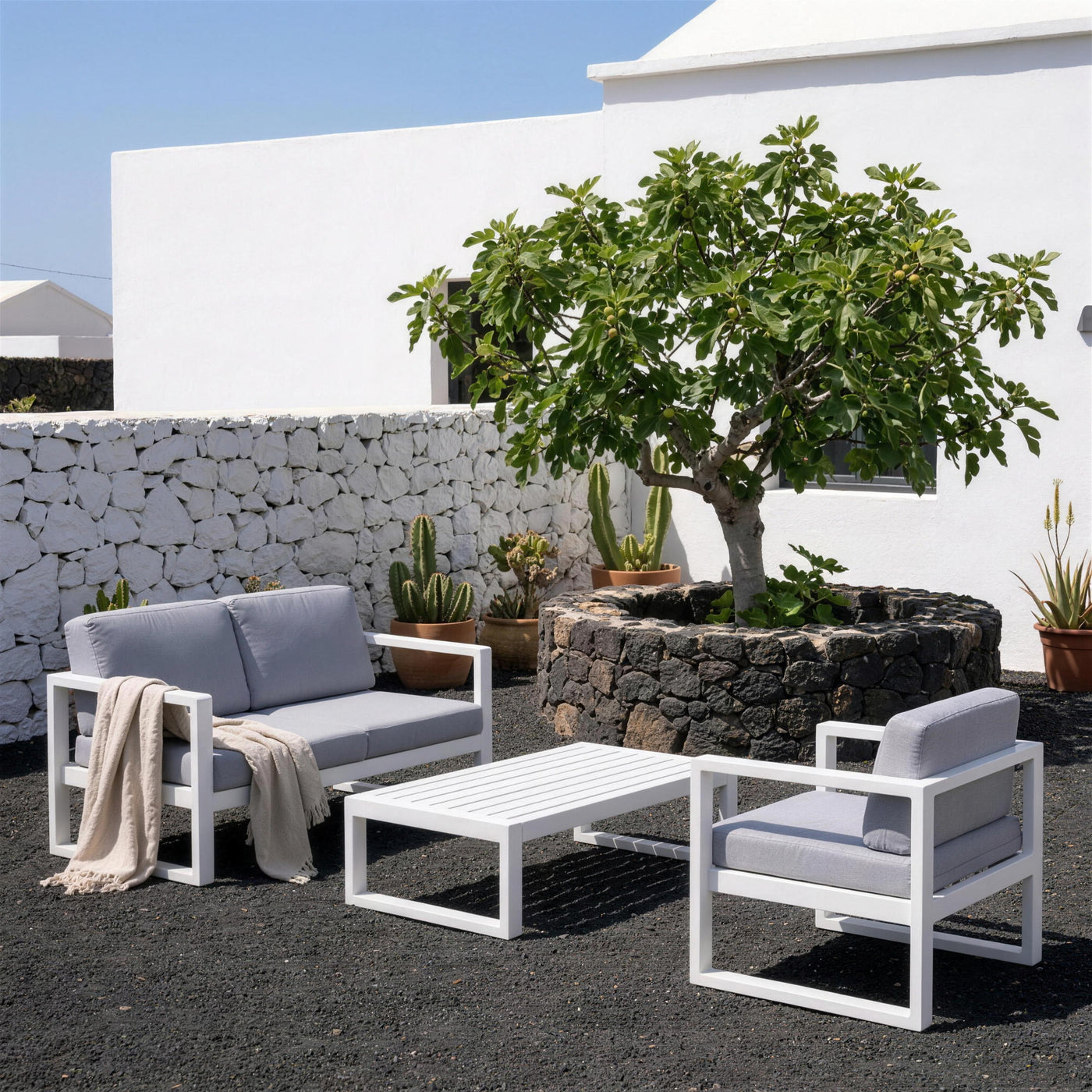 Conjunto Odin-7 blanco en el patio de una casa de Lanzarote, sobre suelo de picon negro, con muro de piedra volcanica, higuera en su zoco y cactus