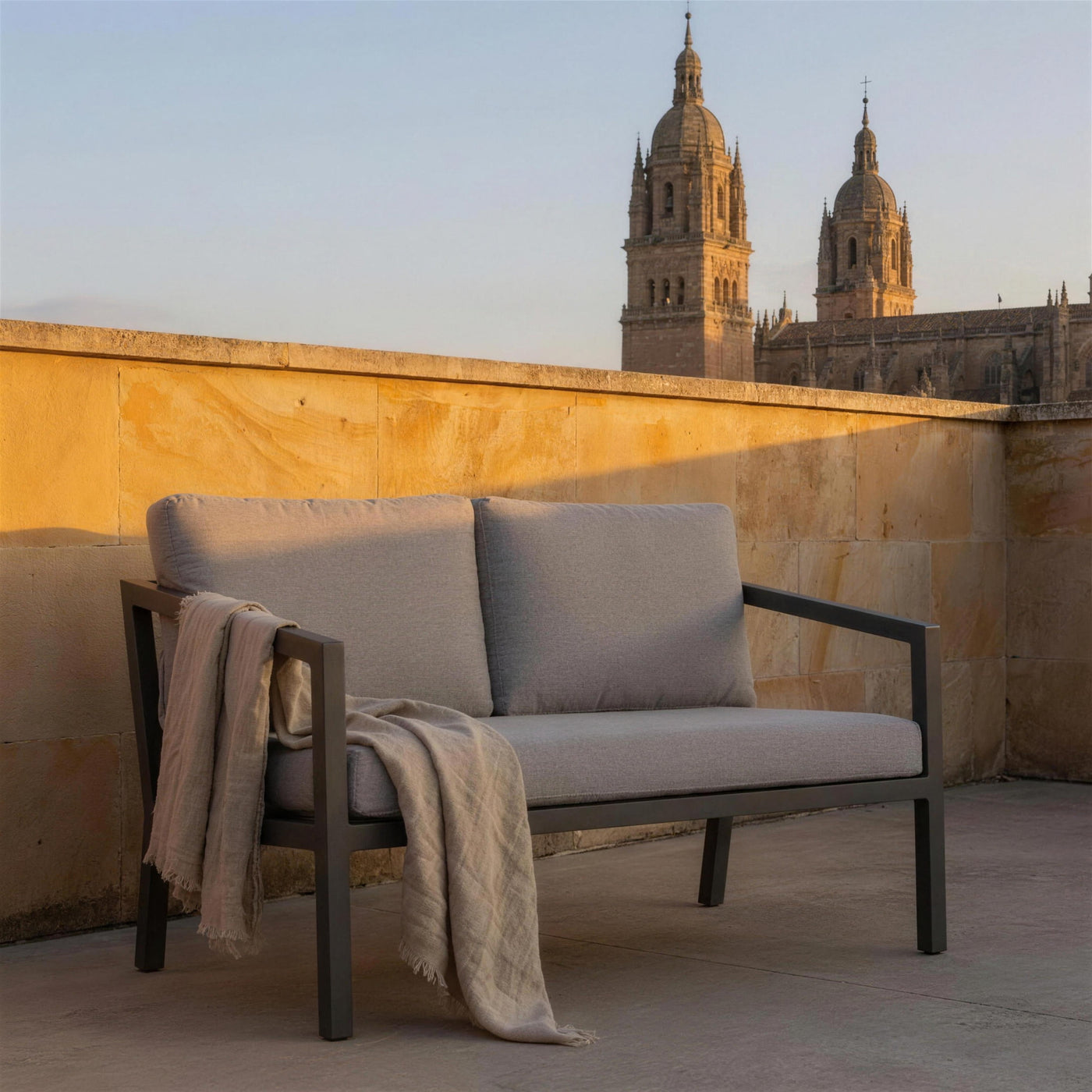 Sofa Acapulco de 2 plazas en una azotea de Salamanca, sobre piedra dorada de Villamayor y con las torres de la catedral al atardecer, con una manta de lino sobre el brazo