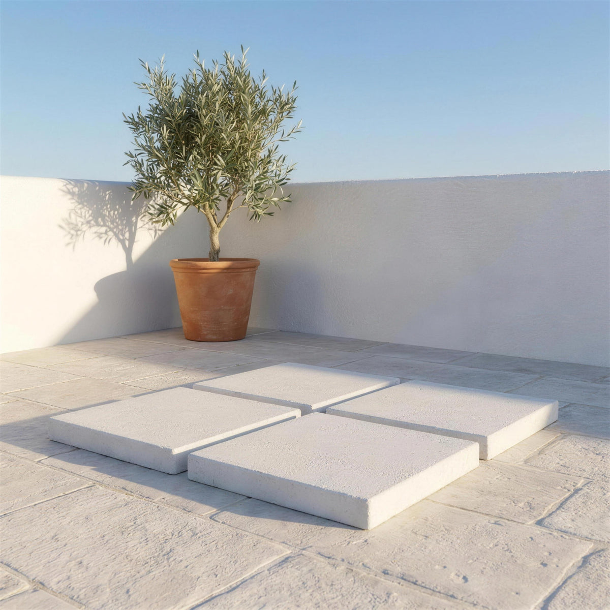 Las cuatro losas de lastre colocadas sobre el suelo de piedra de una terraza encalada, con un olivo en tinaja y un muro blanco al fondo