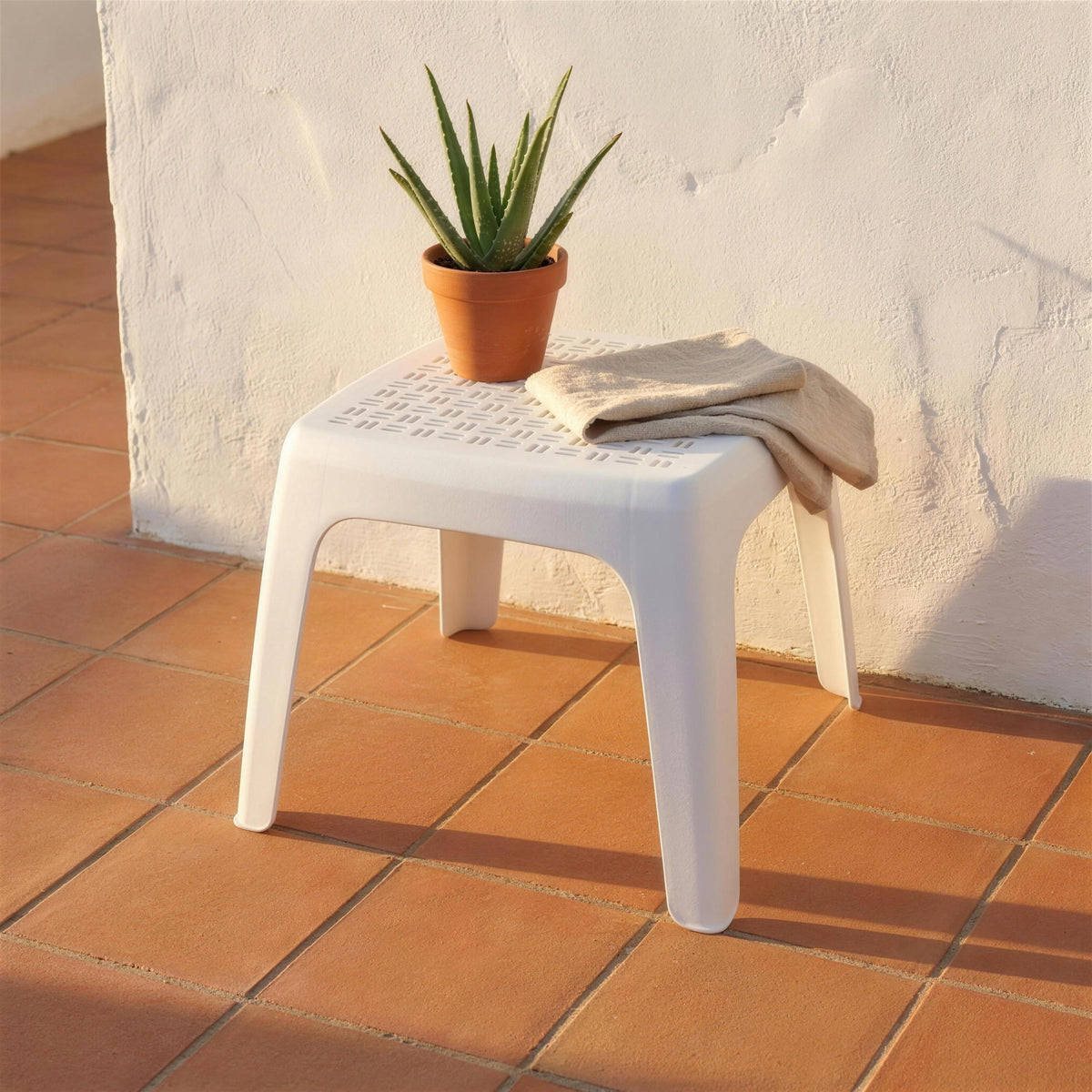 Mesa auxiliar blanca de rejilla, sola en una terraza de barro cocido contra un muro encalado, con un aloe en maceta encima