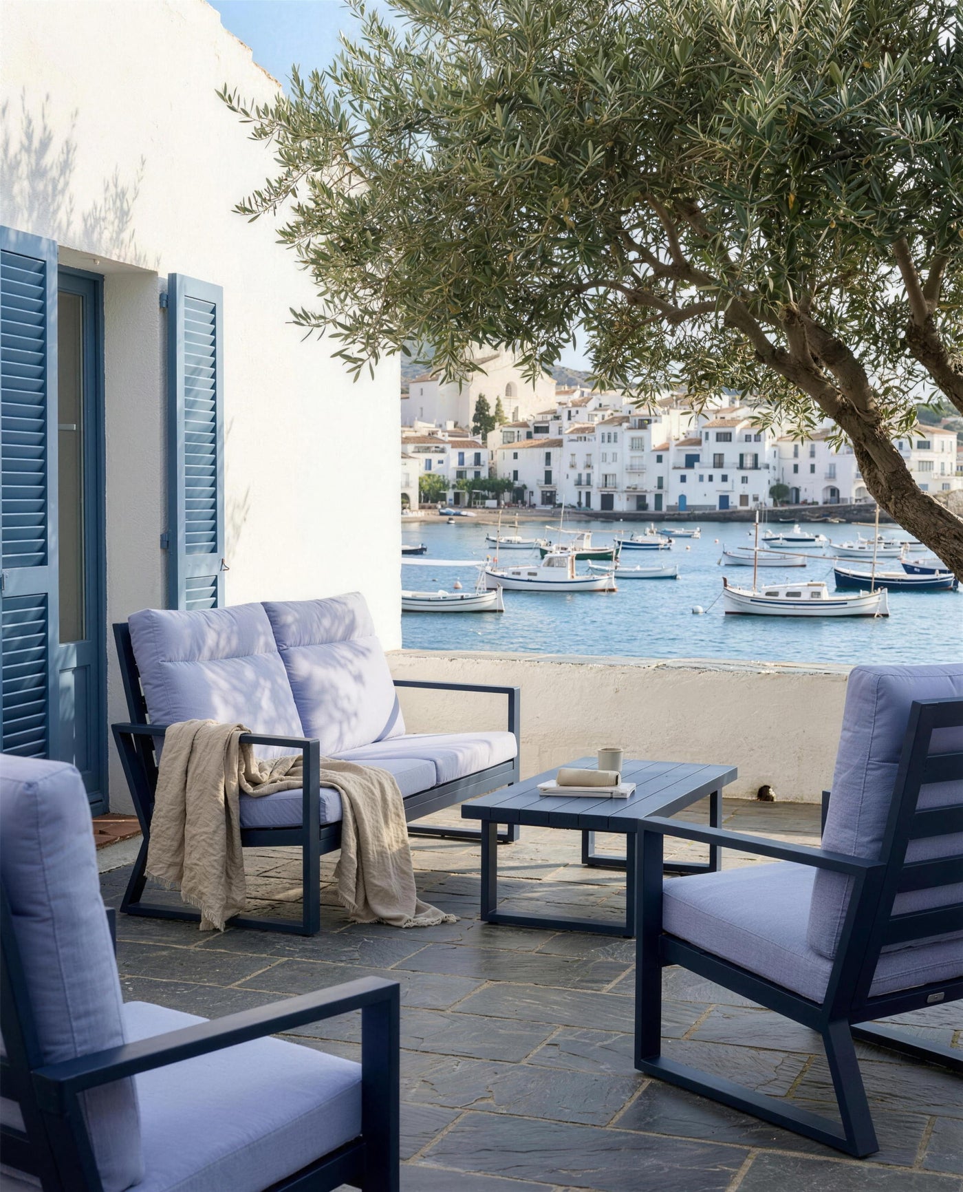 Conjunto Bolonia XL con los postigos azules de lamas y la bahia de Cadaques llena de llauts, con una manta de lino sobre el sofa