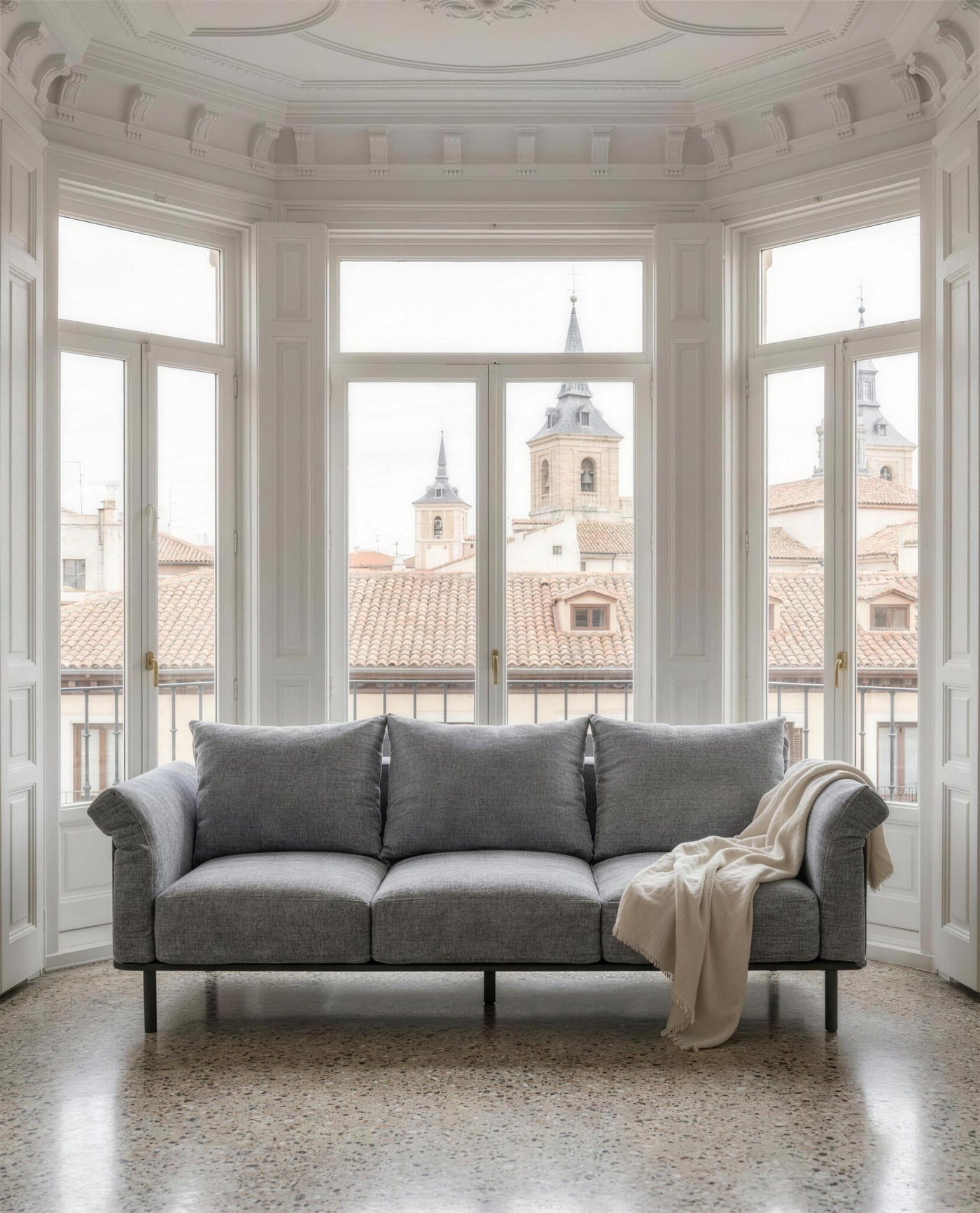 Sofa de 3 plazas gris en el mirador acristalado de un piso madrileno, con molduras, suelo de terrazo y las torres de la ciudad al fondo