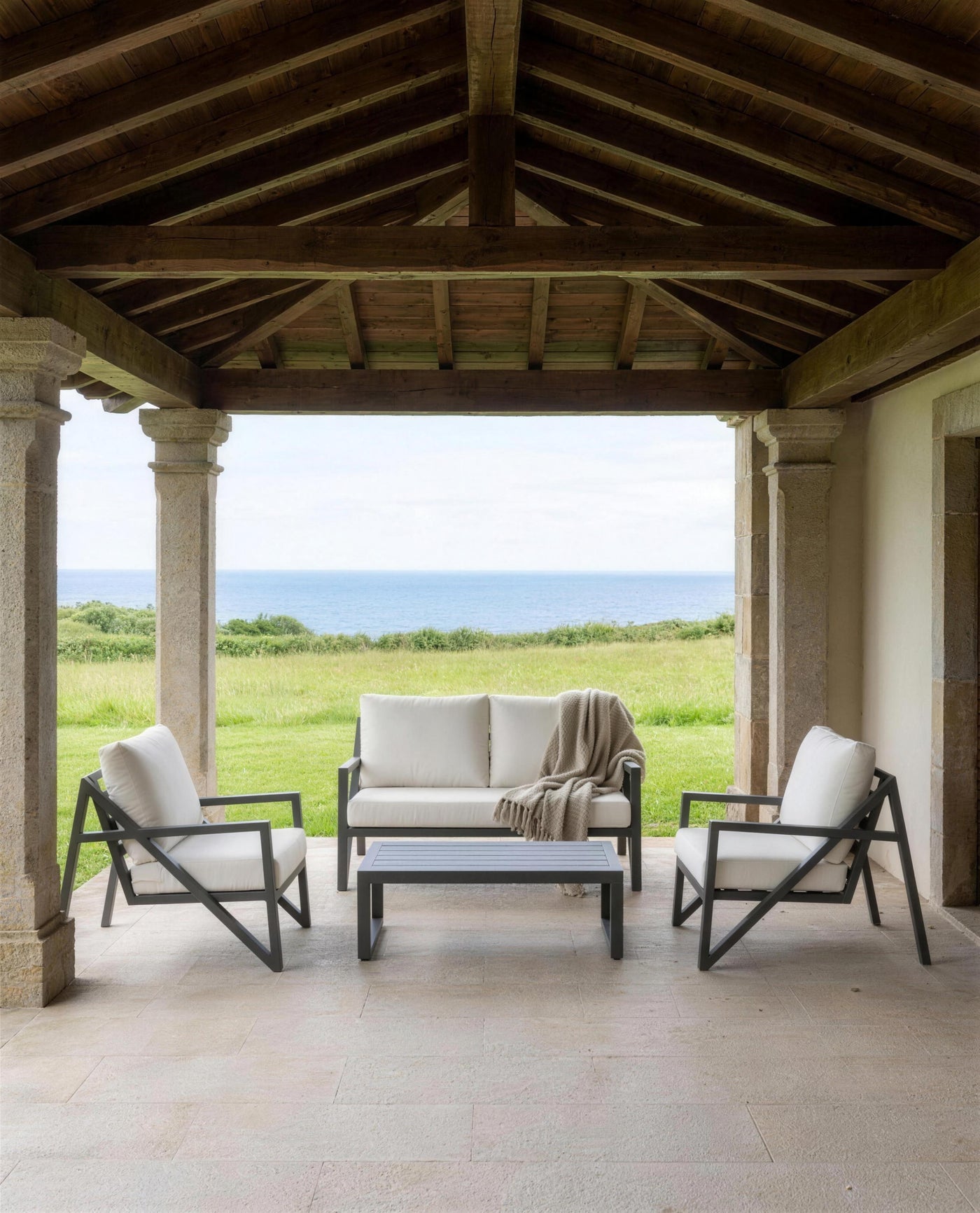 Sofá y sillones de exterior en el porche de columnas de piedra de una casona asturiana, abierto al prado y al mar