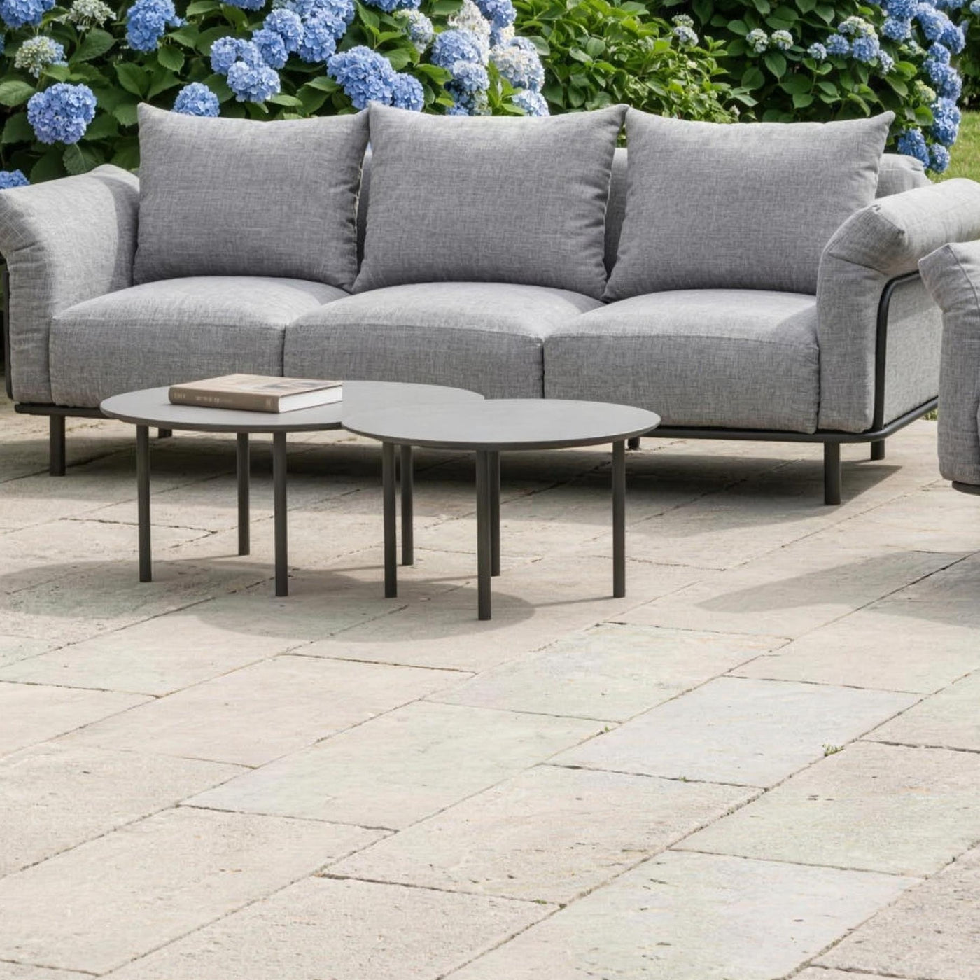 Las dos mesas nido delante del sofa de 3 plazas en la terraza de losa de piedra, con las hortensias azules de la casona detras