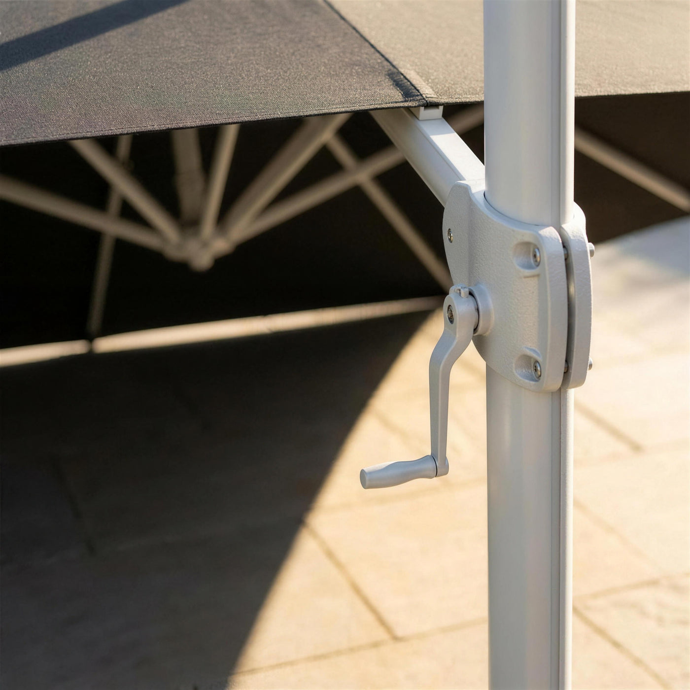 Detalle de la manivela de apertura y el herraje del mástil de aluminio blanco, con la sombra del parasol cruzando el suelo