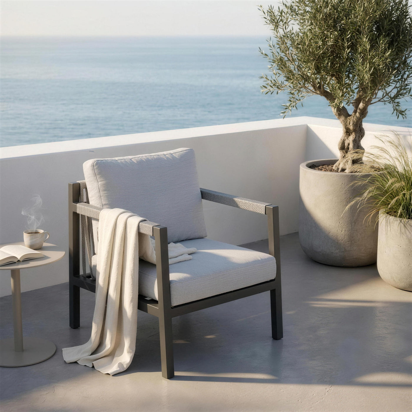 Sillón de exterior antracita en terraza de diseño con suelo de microcemento y olivo, luz de mañana junto al mar