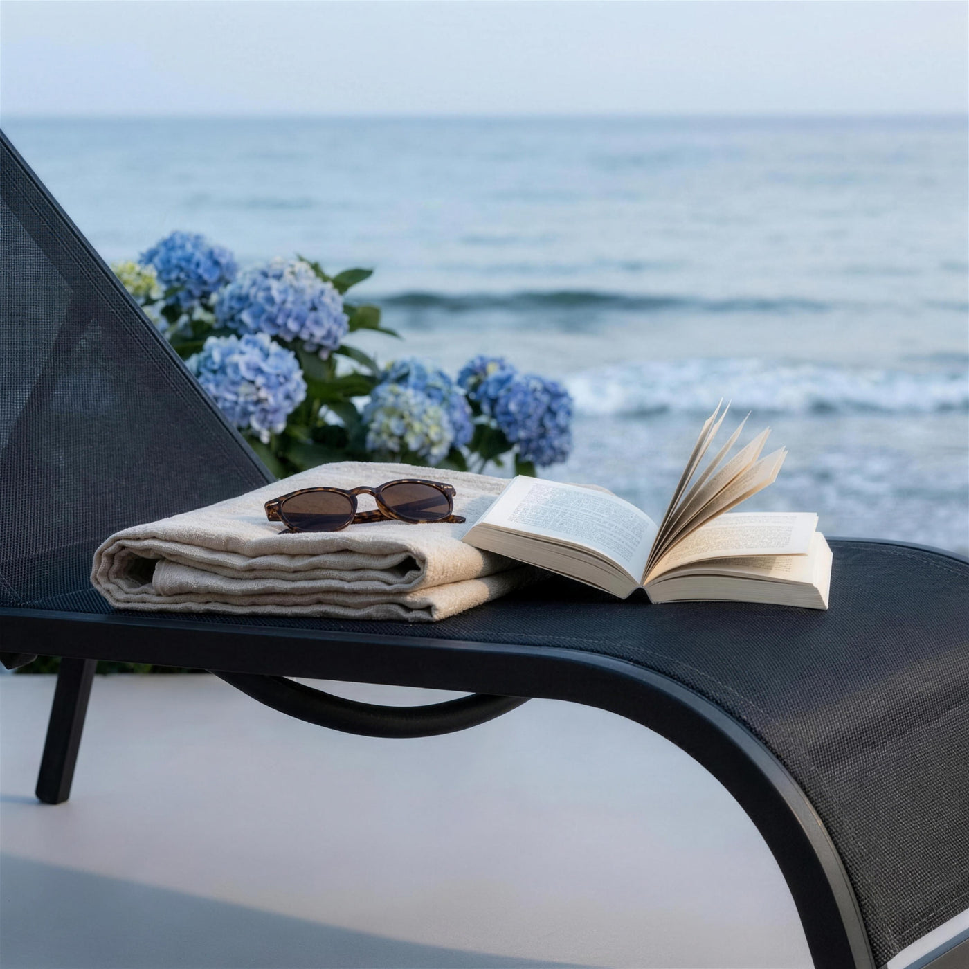 Toalla de lino, gafas de sol y un libro sobre la tumbona Hevea frente al mar