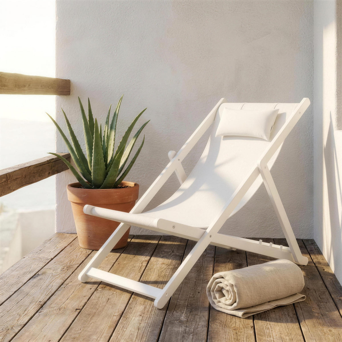 Mini tumbona blanca plegable, sola en un balcon de madera junto a un muro encalado y un aloe en maceta, con una toalla de lino enrollada
