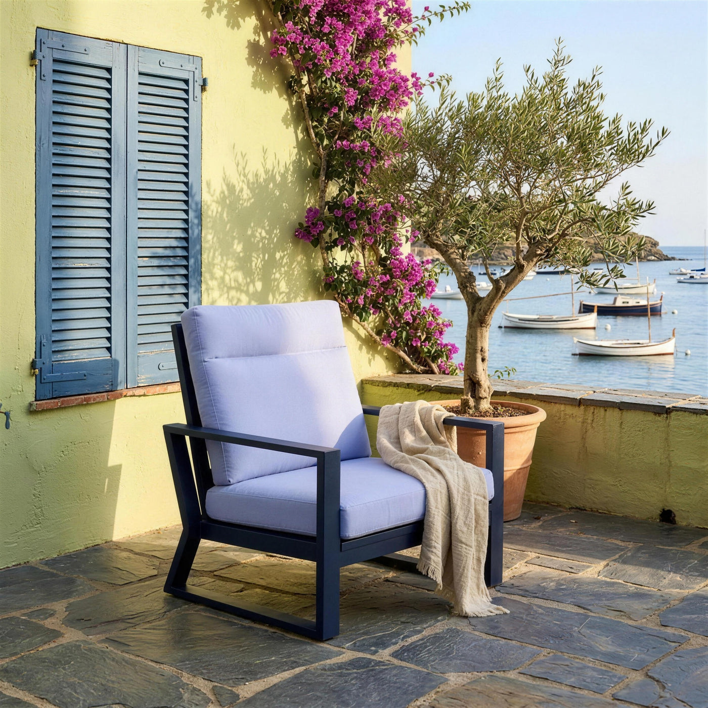 Sillon Bolonia XL en una terraza de Cadaques, sobre laja de pizarra, junto a un olivo en tinaja y con los llauts fondeados en la bahia