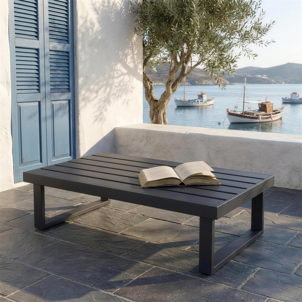 Mesa de centro Bolonia sola en una terraza de Cadaques, sobre laja de pizarra, con muro encalado, postigos de lamas azules, un olivo y la bahia con los llauts al fondo