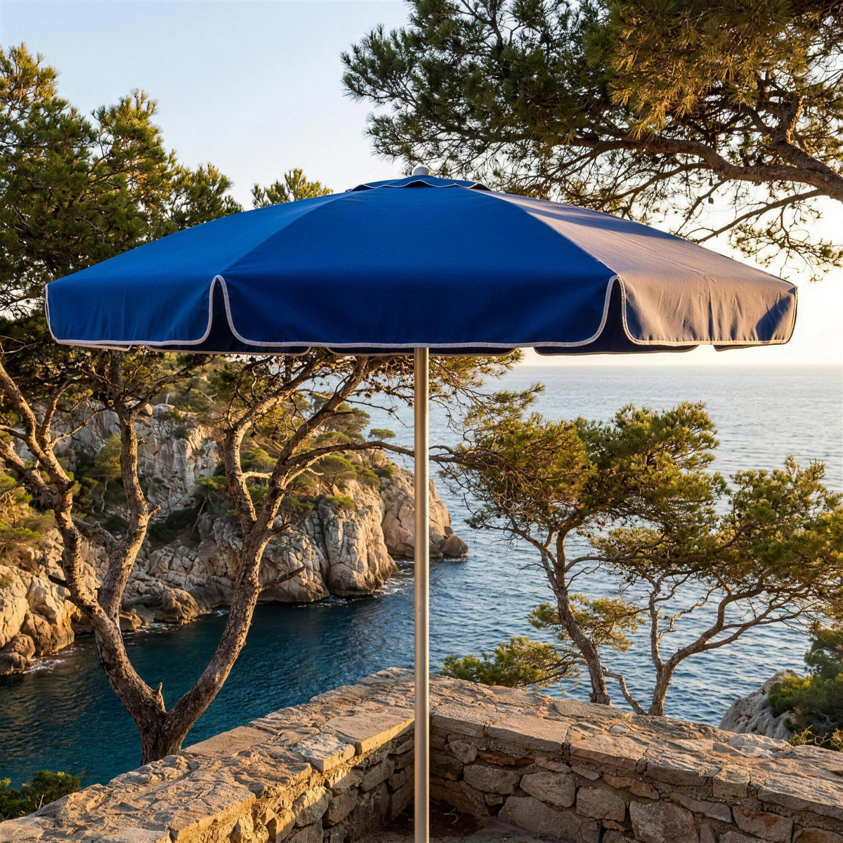 Parasol redondo azul abierto en una terraza de piedra sobre una cala mediterranea con pinos. El pie se vende por separado