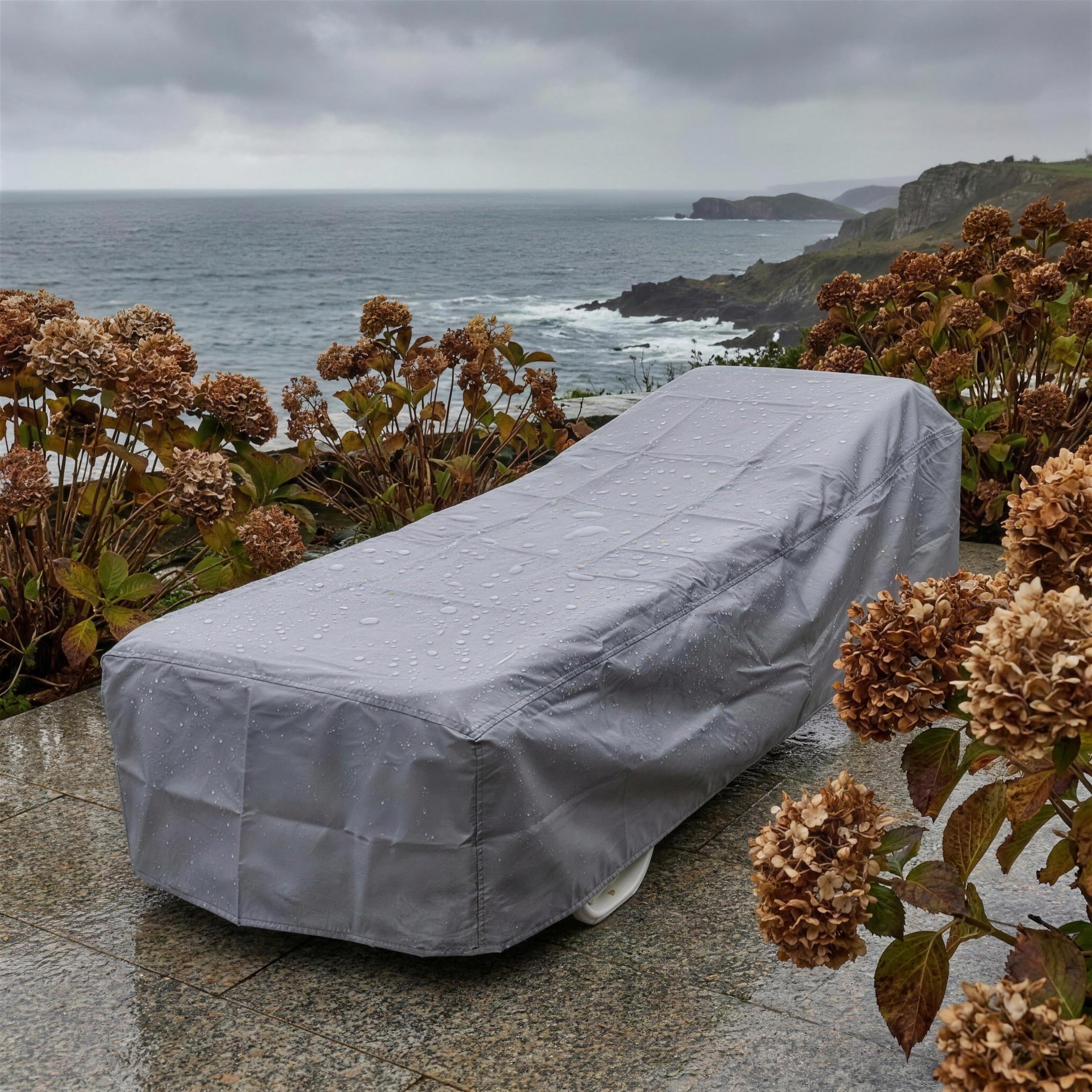 Tumbona cubierta con su funda protectora gris en una terraza de granito del Cantabrico en una manana de otono, con las hortensias pasadas y el mar gris al fondo