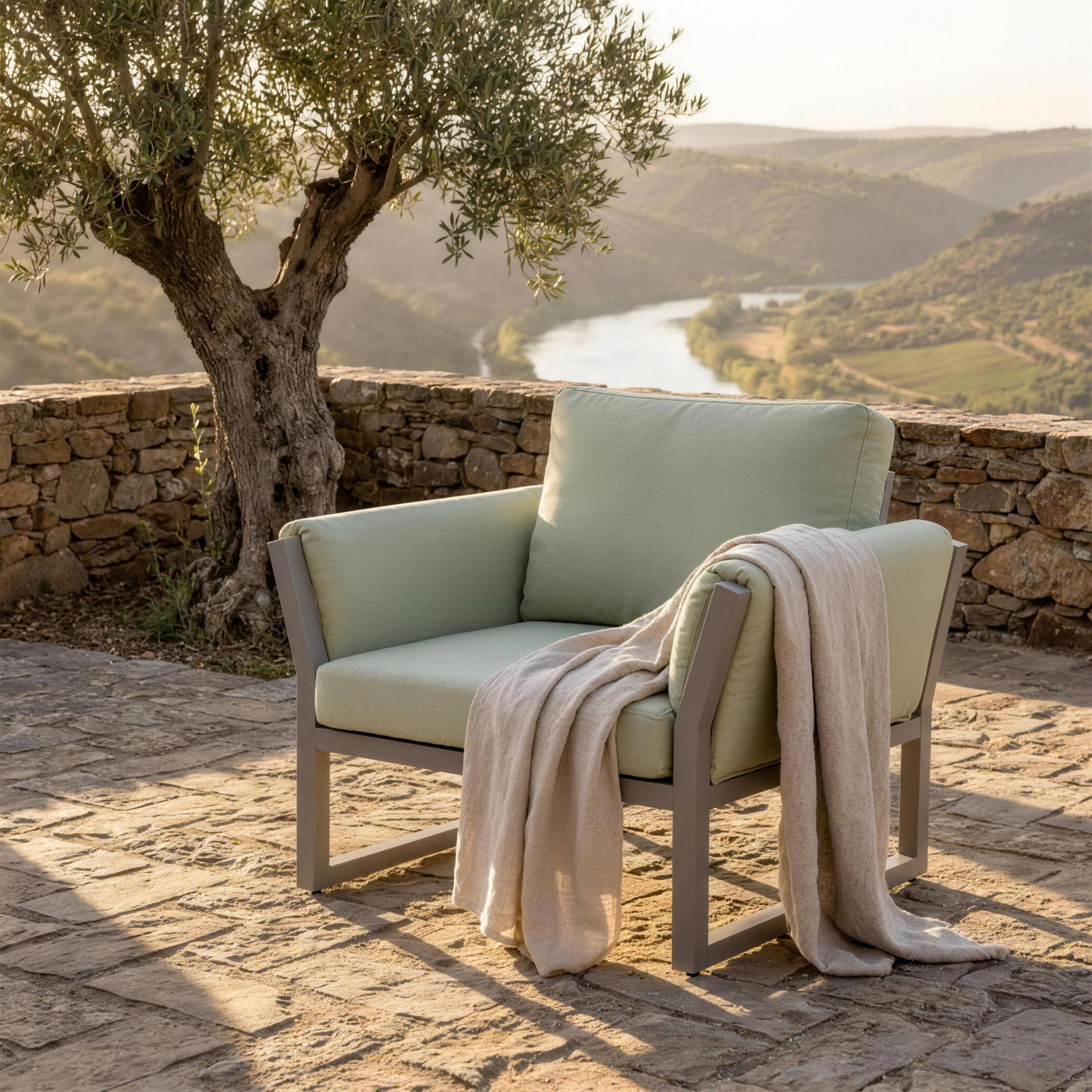 Sillon Cupra en la terraza de un cigarral toledano, junto a un olivo viejo y el muro de mamposteria, con el Tajo y los viñedos al fondo y una manta de lino sobre el brazo