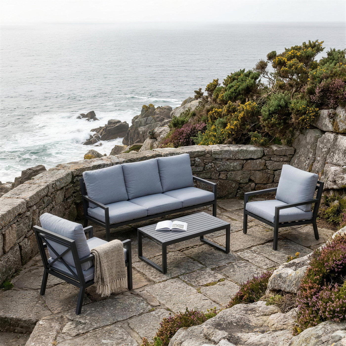 Set de jardín antracita en una terraza de granito de la Costa da Morte, con tojo en flor, brezo y el Atlántico rompiendo abajo