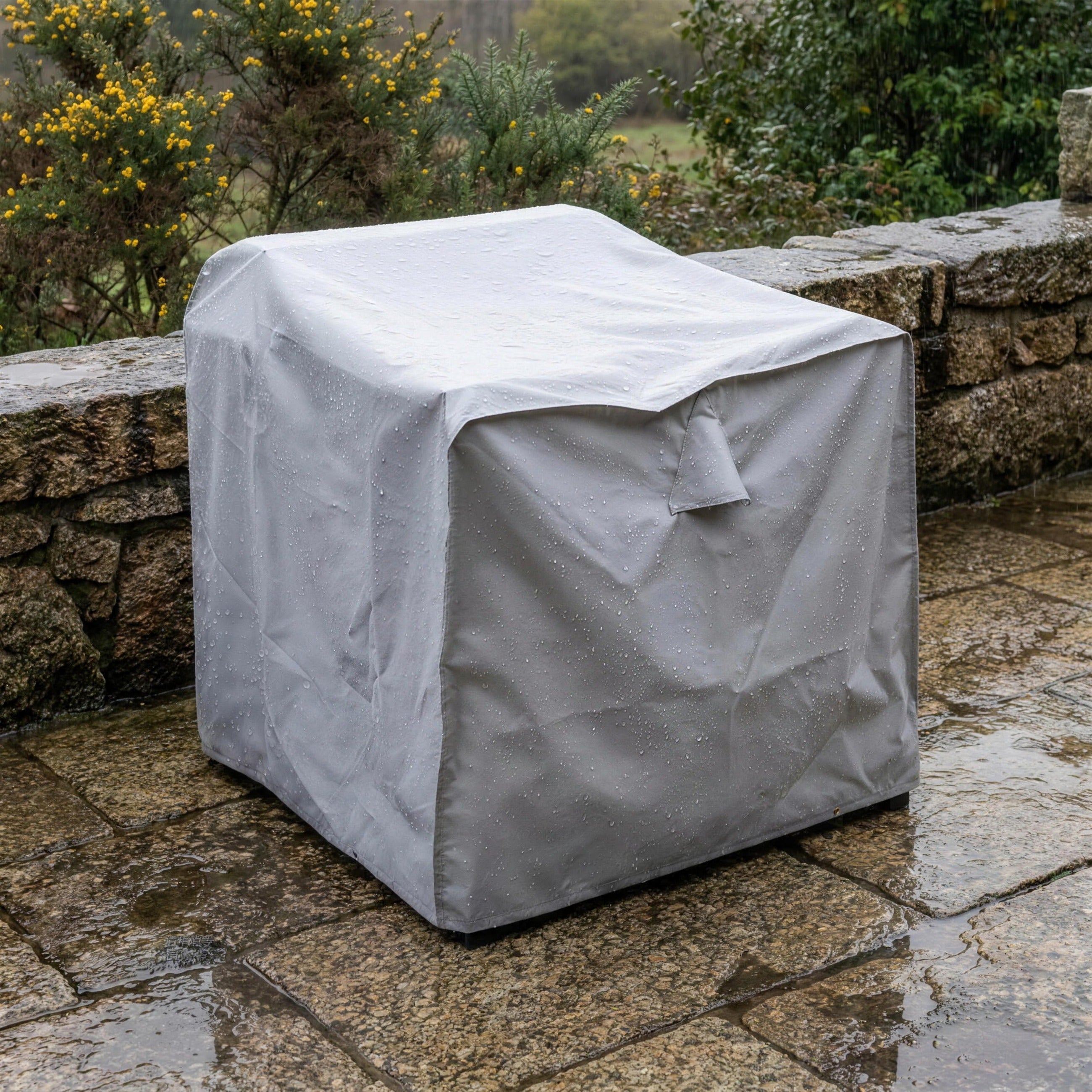 Sillon cubierto con su funda protectora gris en una terraza de granito gallega en una manana de lluvia, con el tojo en flor y las gotas perlando la tela