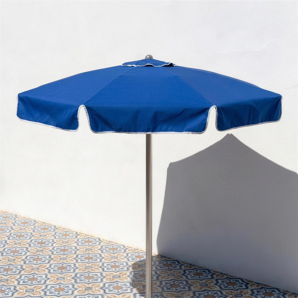 Parasol redondo azul abierto sobre una terraza de baldosa hidraulica junto a un muro encalado, con sombra limpia de mediodia. El pie se vende por separado