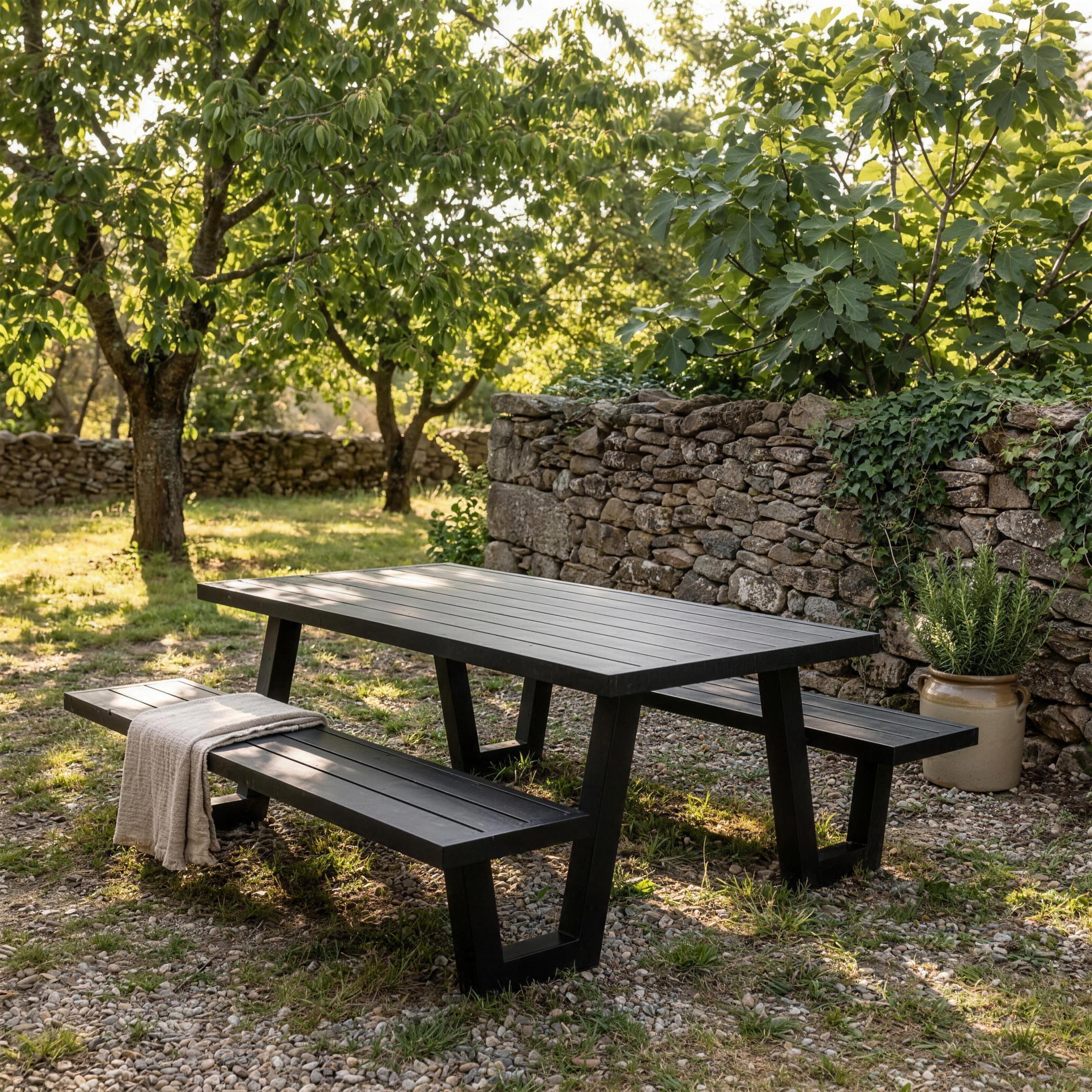 Banco con mesa Yulien en el huerto de una casa de campo extremena, junto a un muro de piedra seca, con cerezos e higuera y romero en una tinaja