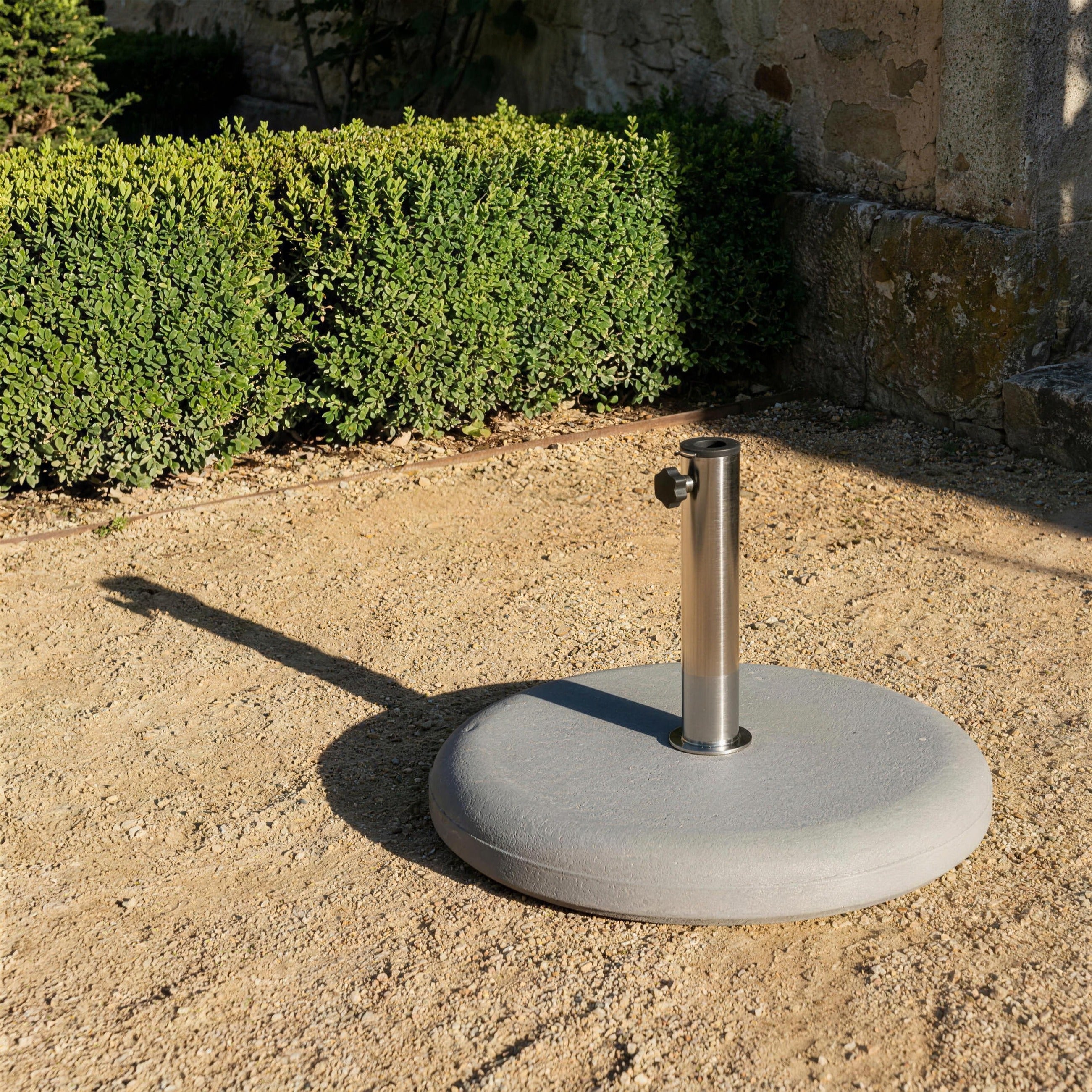 Base de parasol de hormigon sola sobre la grava de un jardin, junto a un seto de boj y un muro de piedra, con su sombra marcada al sol