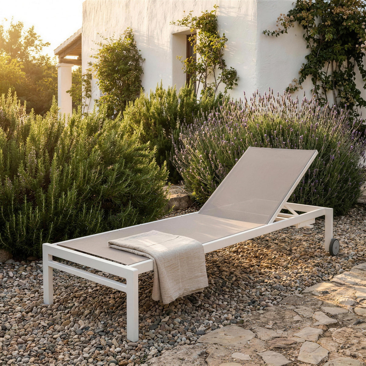 Tumbona de exterior blanca con tejido tortola, sola en un jardin mediterraneo de grava con romero y lavanda junto a una casa encalada