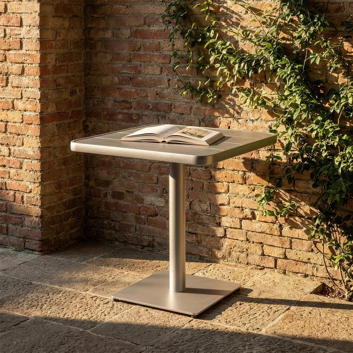 Mesa de exterior de aluminio, sola en un patio contra un muro de ladrillo visto con una enredadera. Se vende solo la mesa, sin sillas