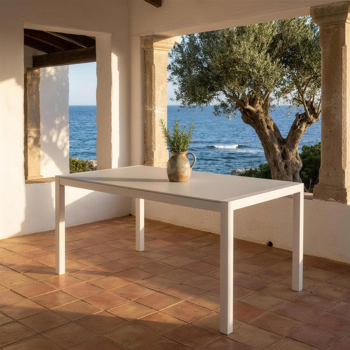 Mesa de comedor de exterior de aluminio blanco sola en el porche encalado de una casa menorquina, con columnas de mares, un olivo y el mar al fondo. Se vende solo la mesa, sin sillas
