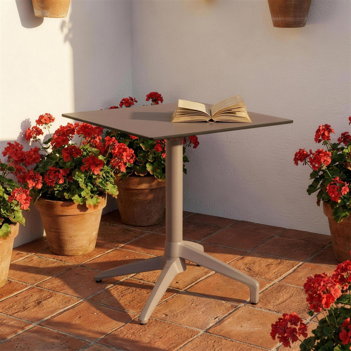 Mesa de exterior Altea con tablero topo, sola en un rincon de patio espanol con suelo de barro cocido, muro encalado y macetas de geranios rojos. Se vende solo la mesa, sin sillas