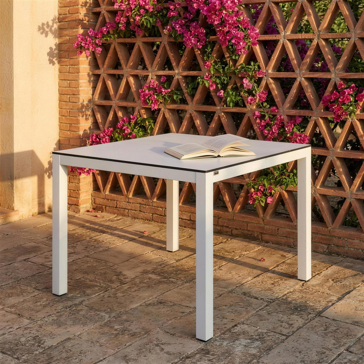 Mesa de exterior blanca, sola en un patio mediterraneo de suelo de piedra junto a una celosia de ladrillo con buganvilla. Se vende solo la mesa, sin sillas
