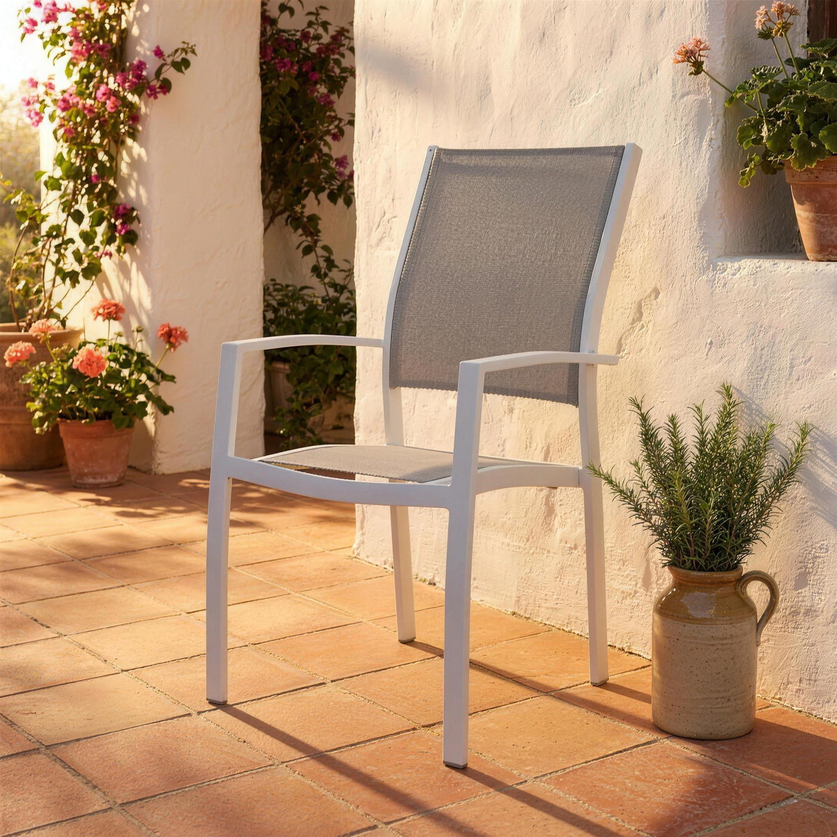 Silla de exterior blanca, sola en un patio espanol de barro cocido junto a un muro encalado y una jarra de gres con romero. Se vende solo la silla