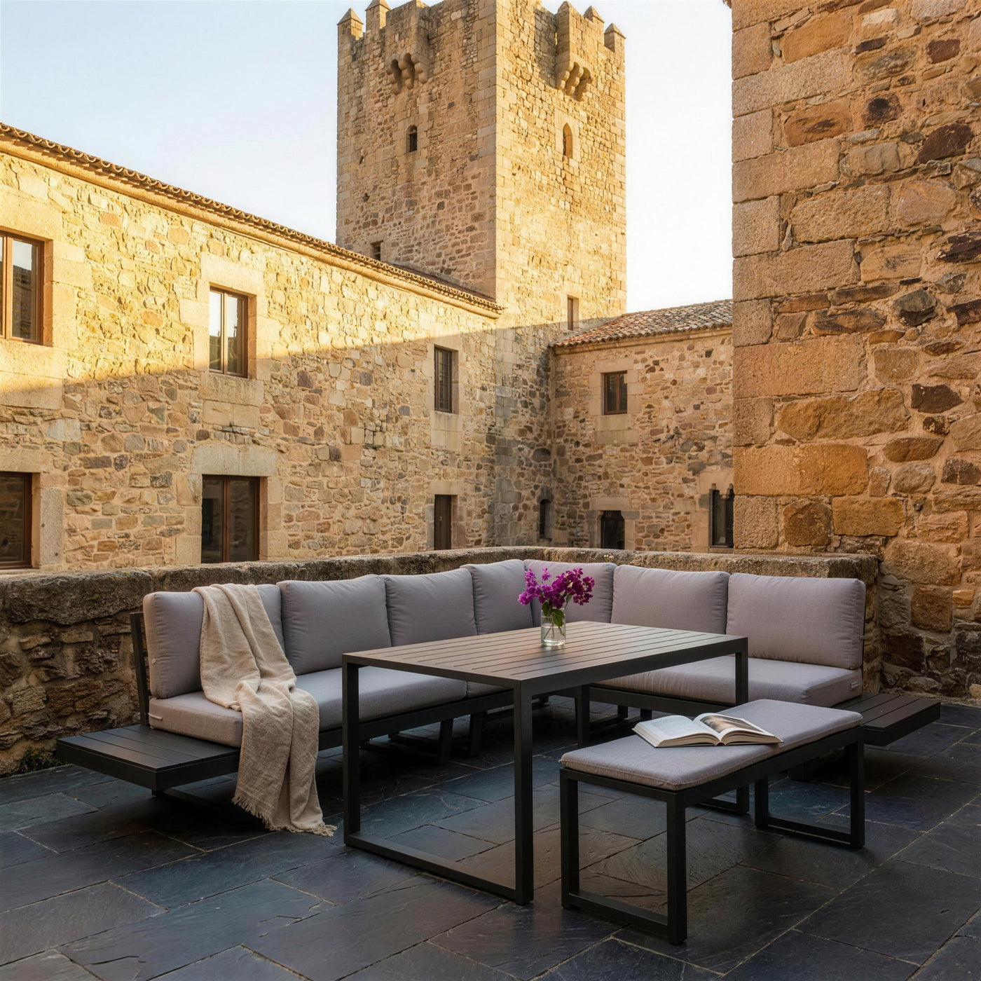 Set rinconera Loira-30 en el patio de una casona de piedra en Caceres, con la torre al fondo, suelo de pizarra, una manta de lino sobre el cojin de esquina y un libro abierto en el banco