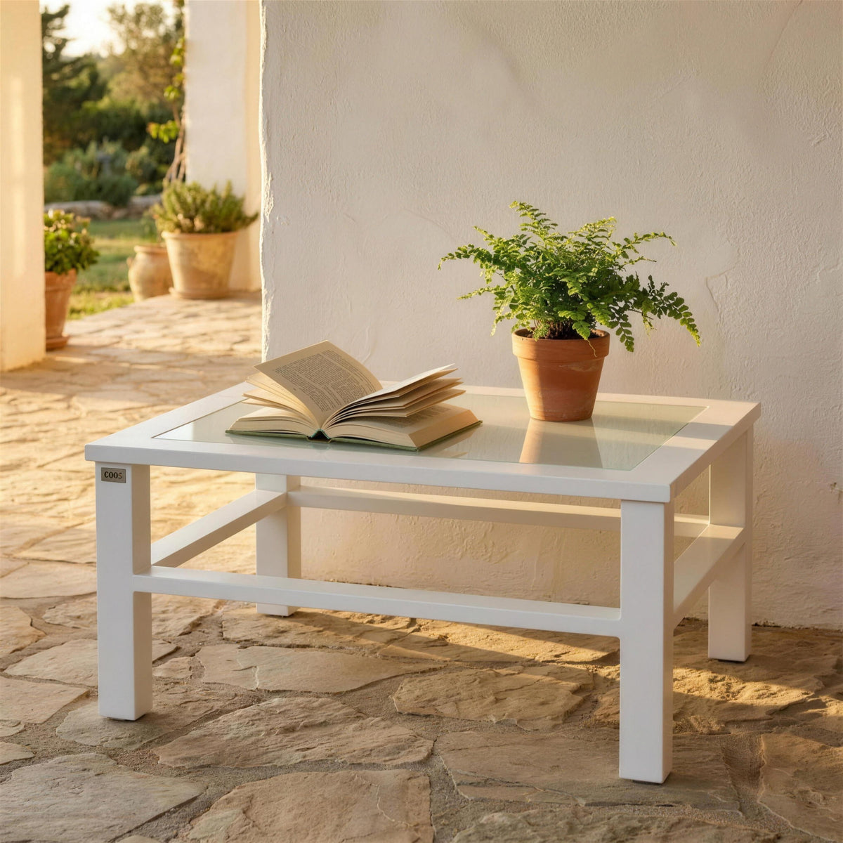 Mesa de centro blanca de exterior, sola en el suelo de piedra de un porche encalado con un libro abierto y un helecho en maceta. Se vende solo la mesa