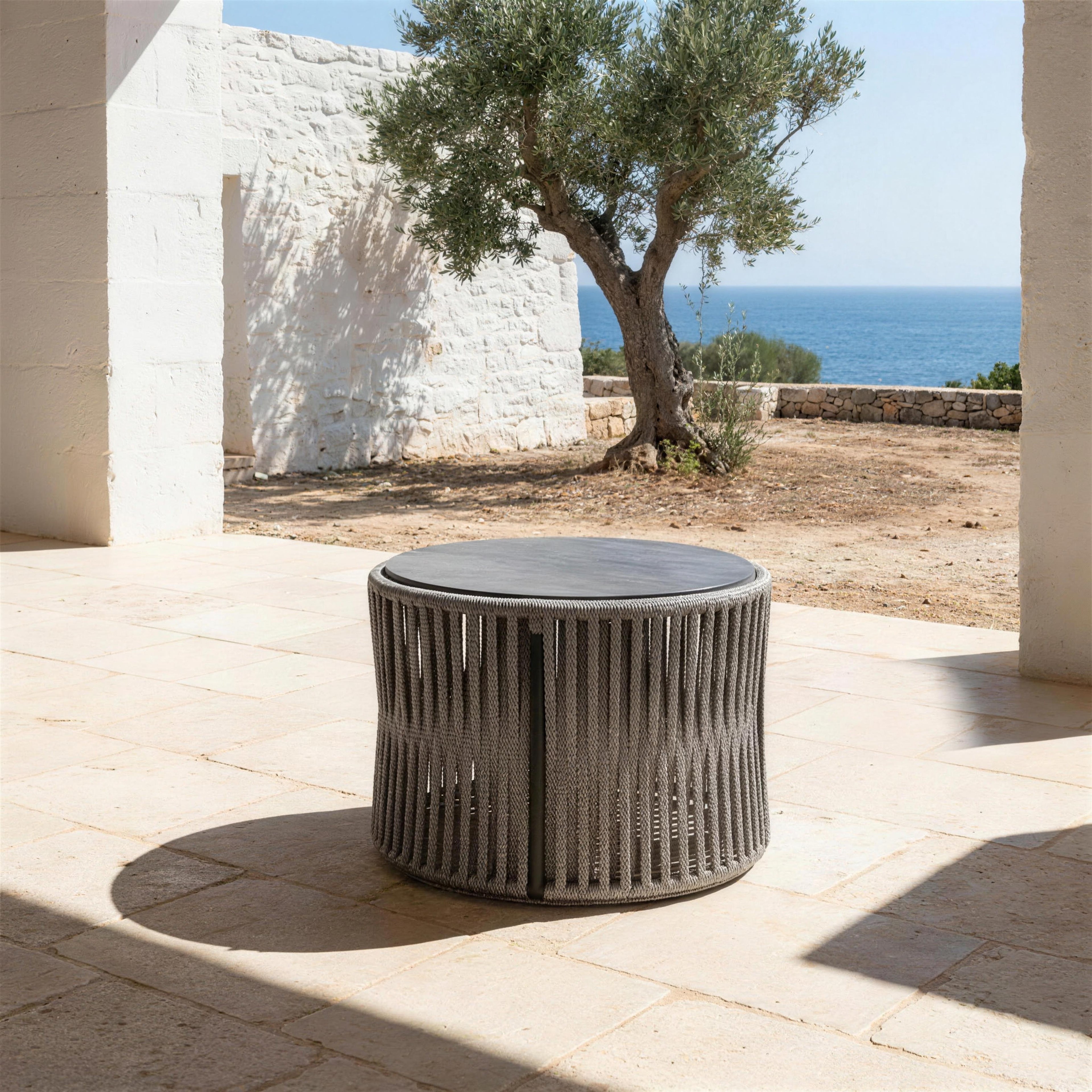 Mesa de centro Yina de cuerda trenzada sobre el suelo de piedra de una porxada menorquina, con el olivo y el mar detras
