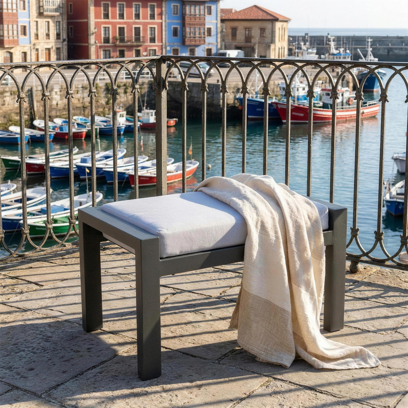 Reposapies Standard XL antracita solo en una terraza de piedra sobre un puerto pesquero asturiano, con reja de forja y las barcas abajo