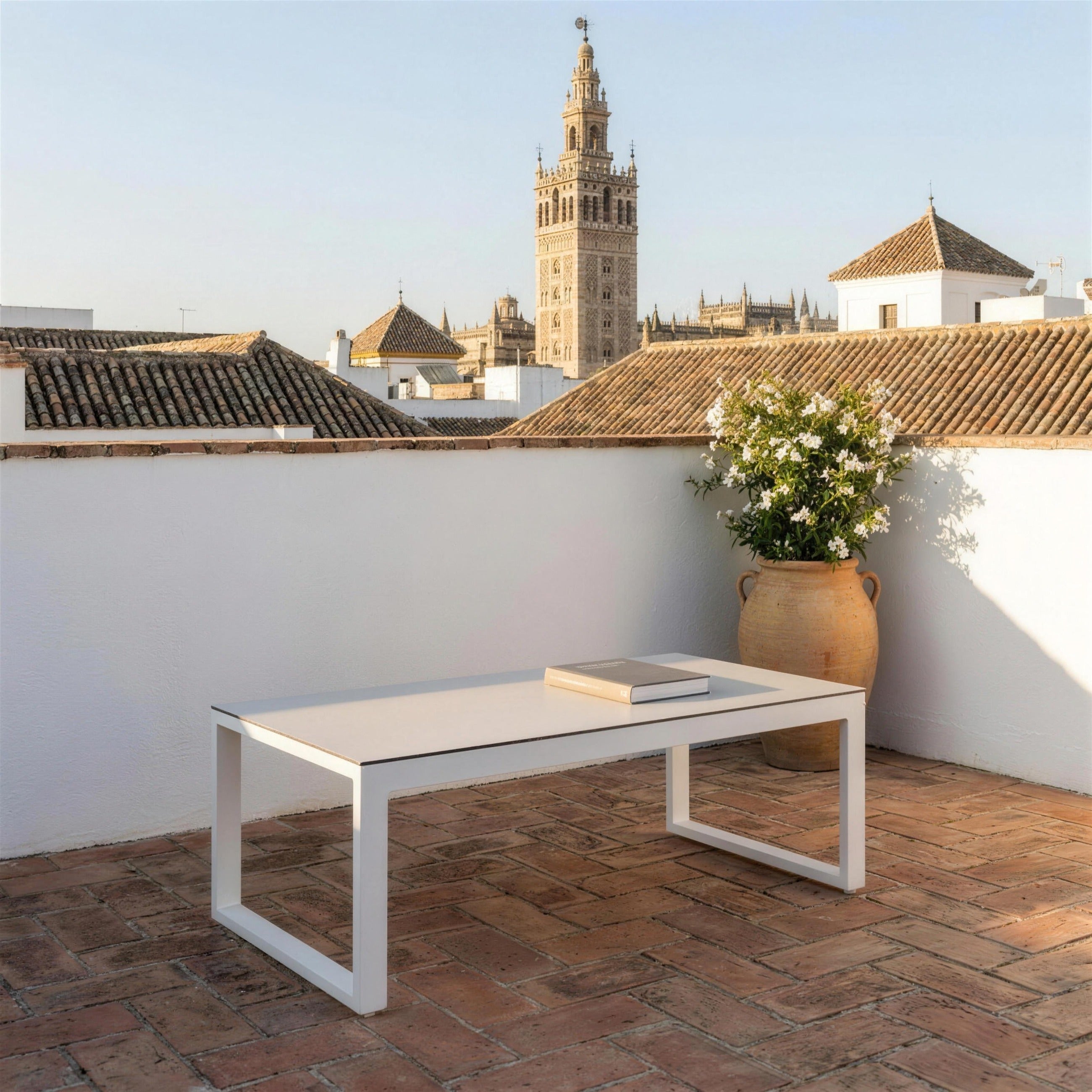 Mesa de centro blanca con tablero HPL sola en una azotea sevillana de suelo de ladrillo, con jazmin en tinaja y la Giralda sobre los tejados