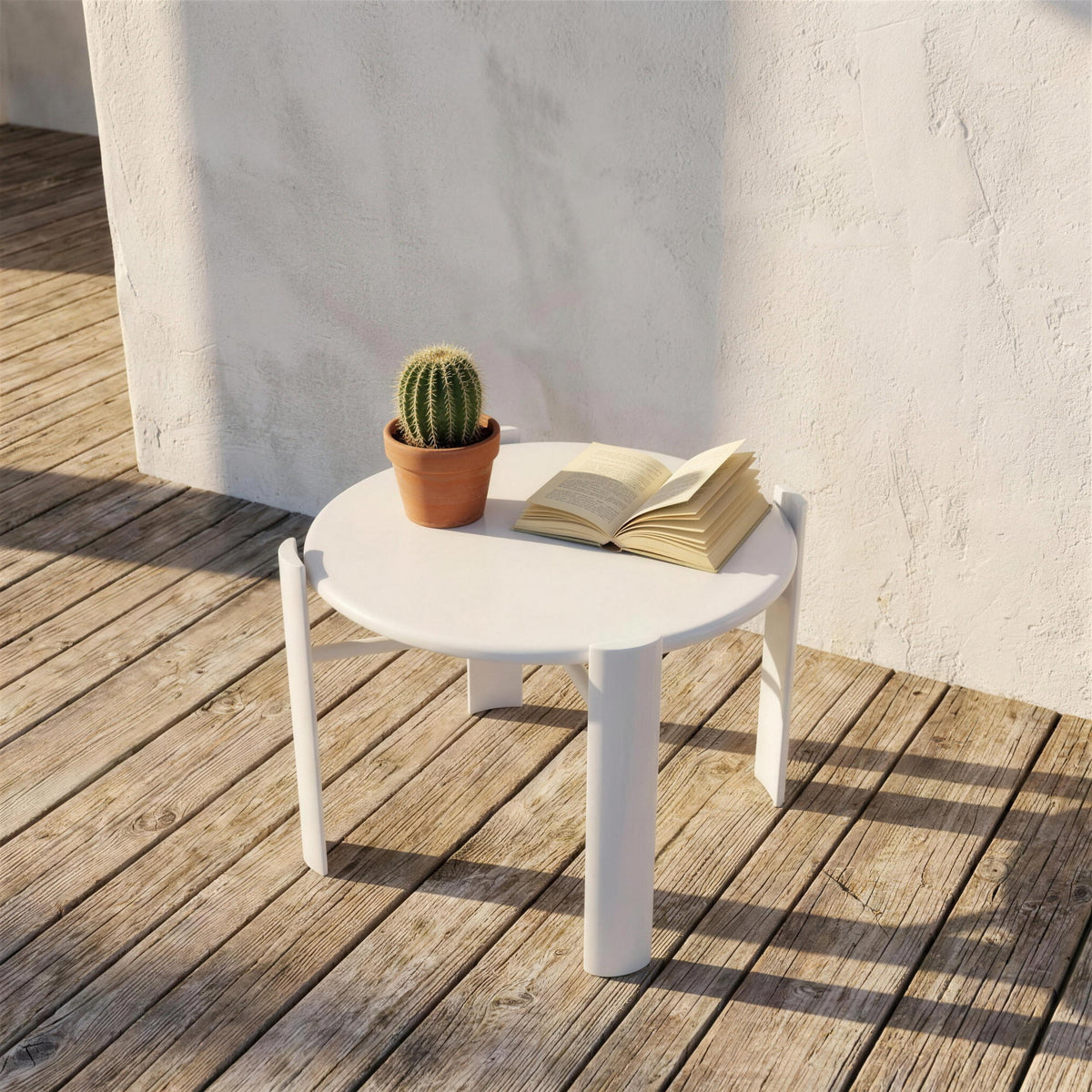 Mesa auxiliar redonda blanca, sola en una tarima de madera junto a un muro encalado, con un cactus en maceta y un libro abierto encima