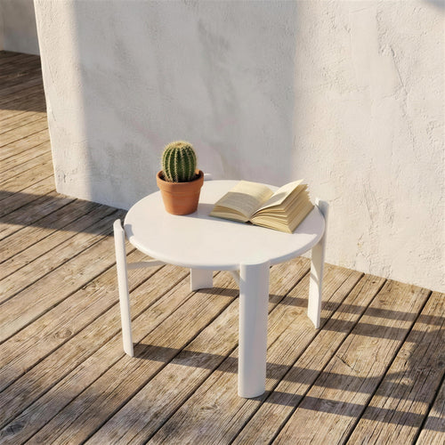 Mesa auxiliar redonda blanca, sola en una tarima de madera junto a un muro encalado, con un cactus en maceta y un libro abierto encima