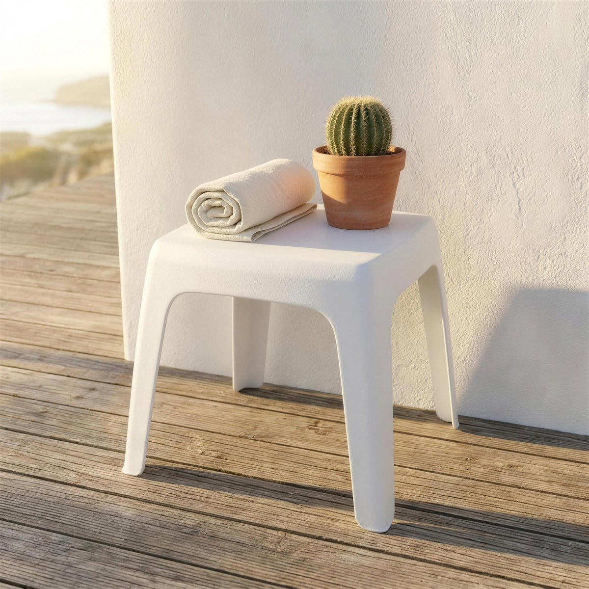 Mesa auxiliar blanca de tablero liso, sola en una terraza de tarima de madera junto a un muro encalado, con una toalla enrollada y un cactus en maceta