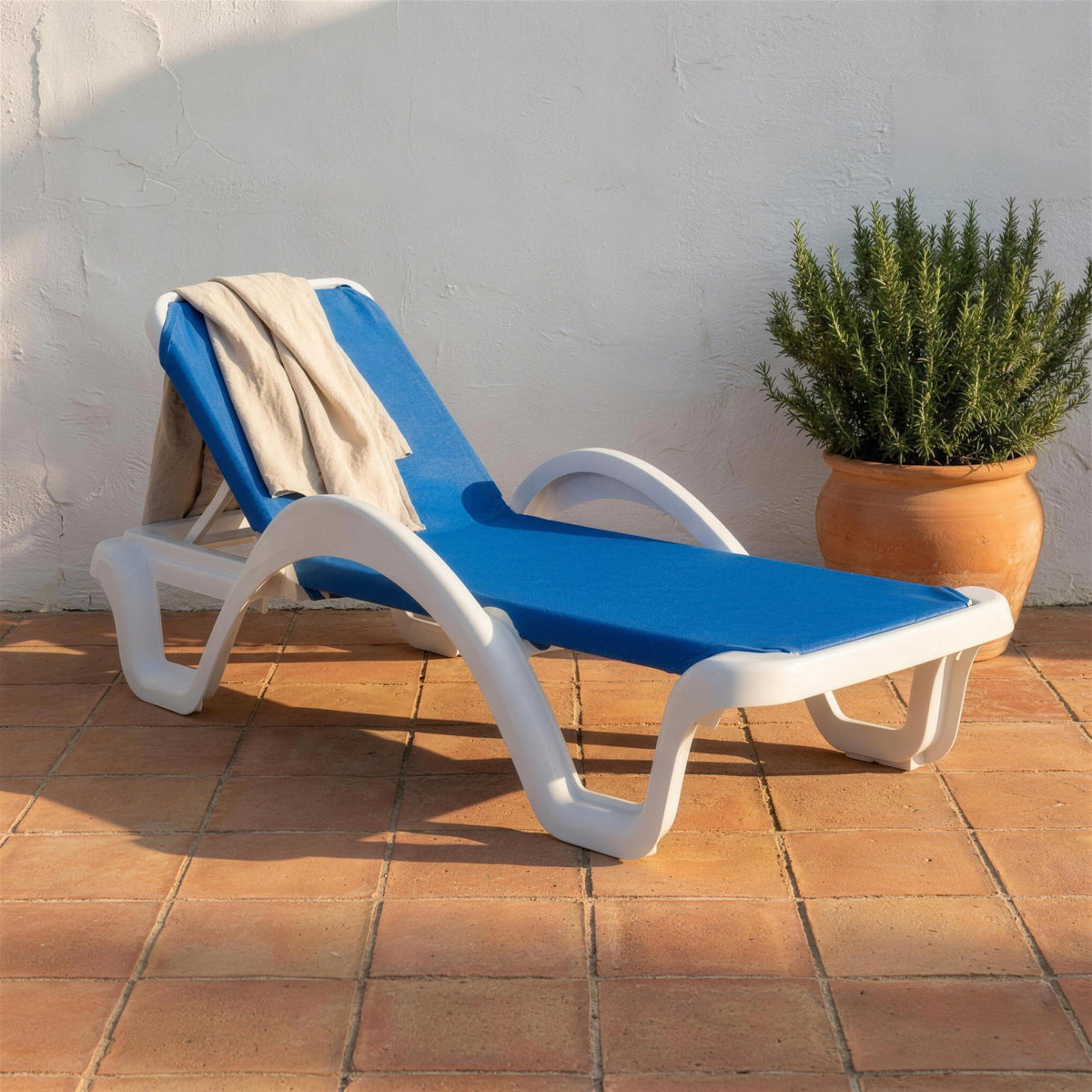 Tumbona de resina blanca con tejido azul, sola en una terraza de barro cocido junto a un muro encalado con romero en maceta y una toalla de lino