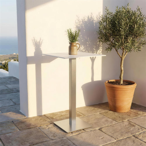 Mesa alta de exterior sola en una terraza mediterranea encalada con suelo de piedra, un olivo en maceta y una jarra de gres con romero encima. Se vende solo la mesa, sin taburetes