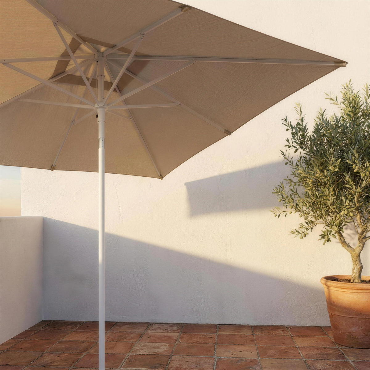Parasol cuadrado color avena abierto sobre una terraza mediterranea de barro cocido, junto a un muro encalado y un olivo en maceta. El pie se vende por separado