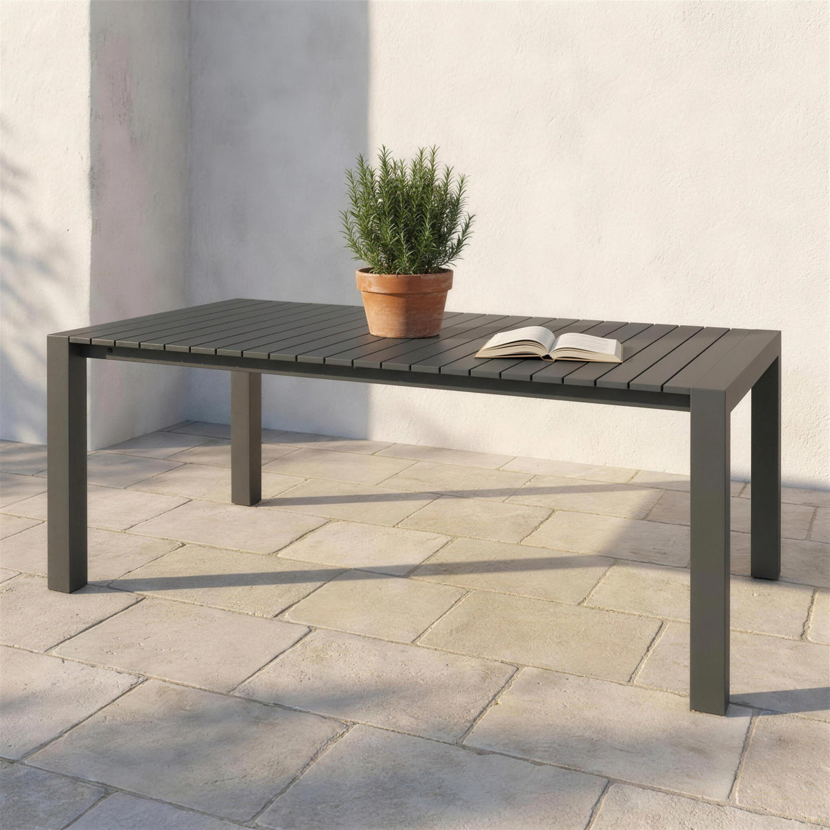 Mesa extensible de exterior antracita con tablero de listones, sola en una terraza de piedra clara junto a un muro encalado y una maceta de romero. Se vende solo la mesa, sin sillas