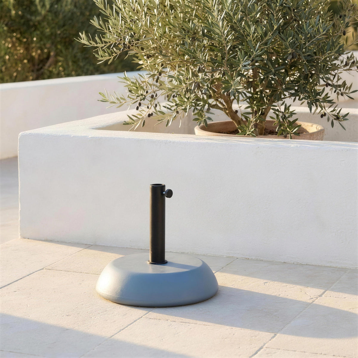 Base de parasol de 25 kg sola sobre el suelo de piedra de una terraza encalada, junto a una jardinera con olivos. Se vende solo la base, sin parasol