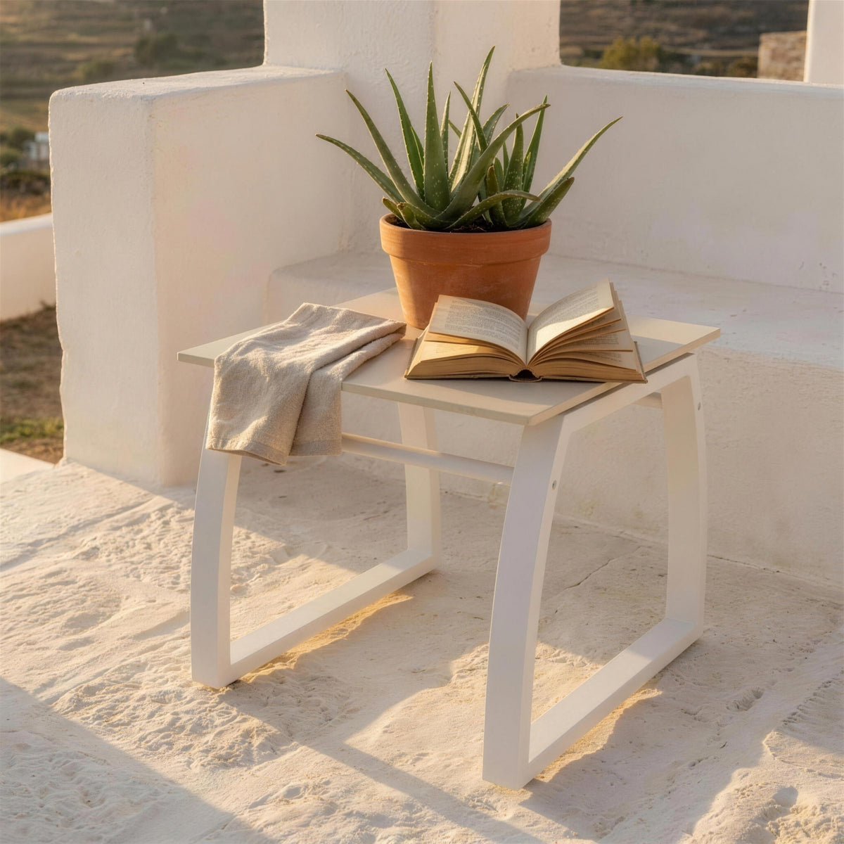Mesa auxiliar blanca de exterior, sola en el suelo de piedra de una terraza encalada junto a un aloe en maceta, con un libro abierto encima