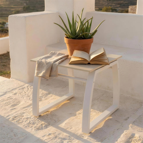 Mesa auxiliar blanca de exterior, sola en el suelo de piedra de una terraza encalada junto a un aloe en maceta, con un libro abierto encima