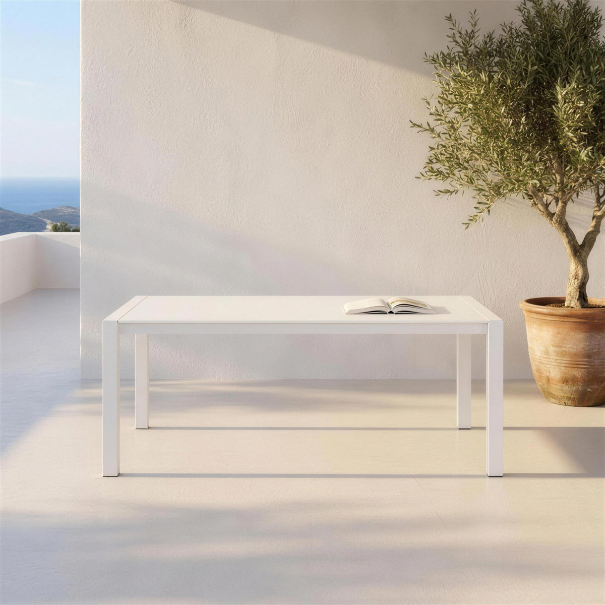 Mesa extensible de exterior blanca, sola en una terraza mediterranea de microcemento junto a un muro encalado y un olivo en maceta de barro. Se vende solo la mesa, sin sillas