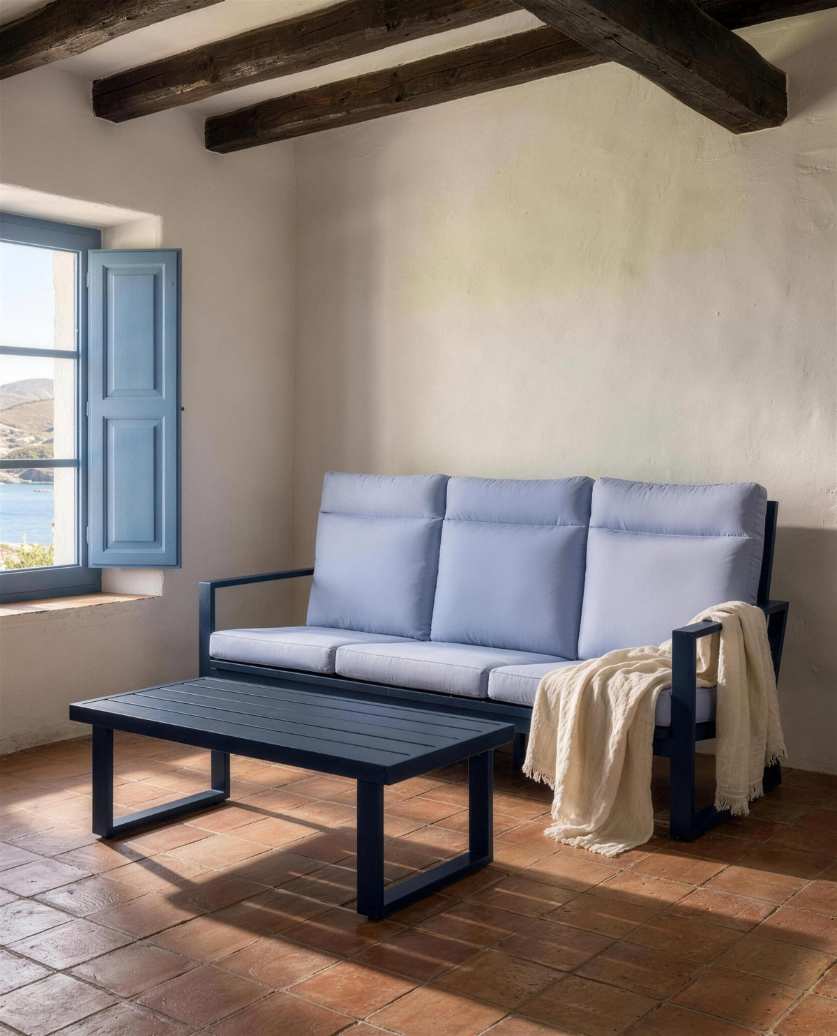 Sofa de 3 plazas y mesa de centro azul marino en una sala encalada de casa de pescadores, con vigas de madera, suelo de barro cocido y la ventana abierta al mar
