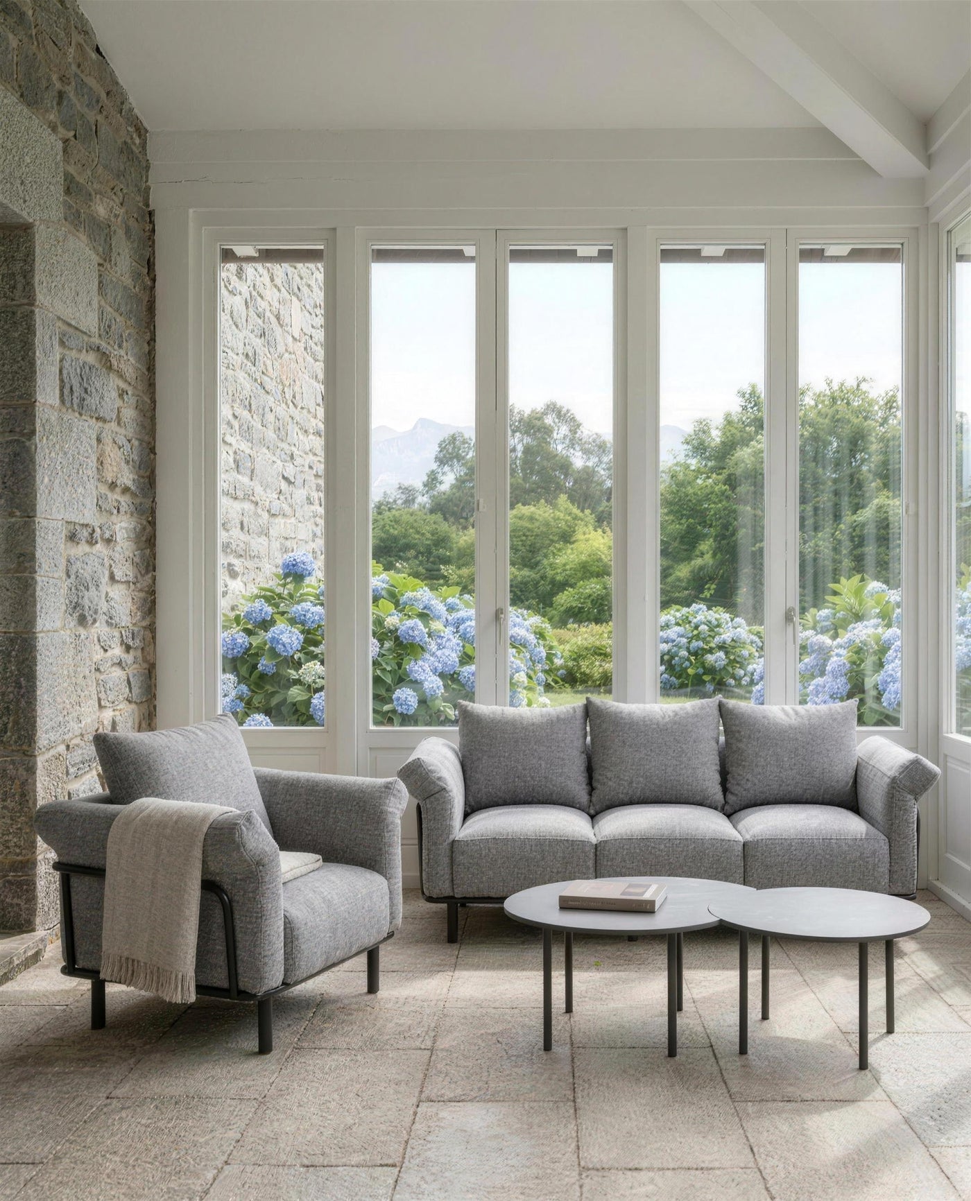 Sofa de 3 plazas y sillon de exterior con las dos mesas nido en la galeria acristalada de una casona de piedra, con las hortensias al otro lado del cristal