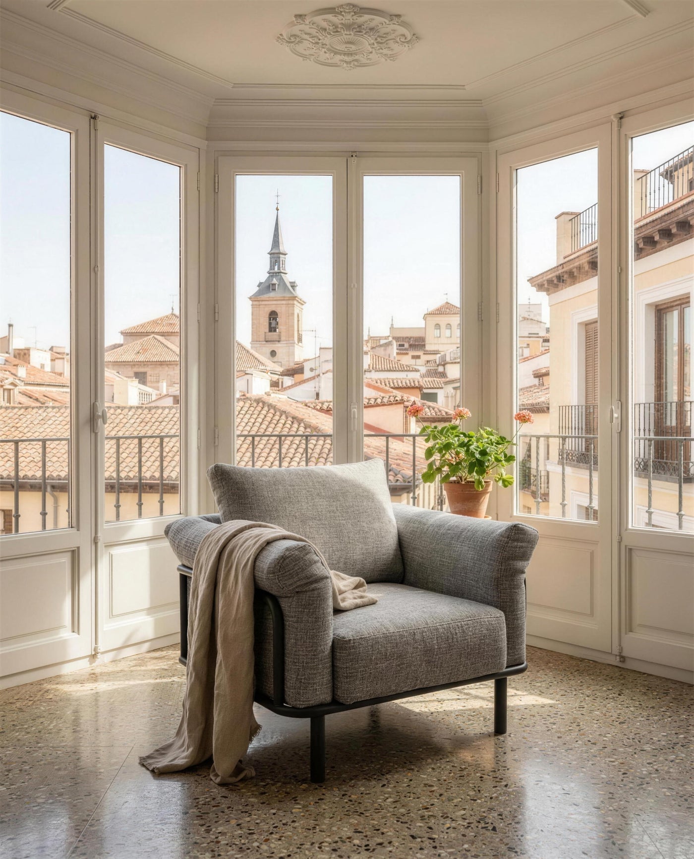 Sillon gris en el mirador acristalado de un piso madrileno, con molduras, suelo de terrazo, un geranio en maceta de barro y los tejados de la ciudad al fondo