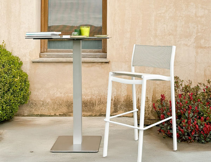 Taburete alto de exterior de aluminio blanco con asiento y respaldo de tejido, modelo Etna, junto a una mesa alta